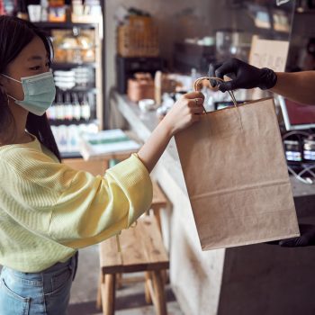 Asian woman wearing protective mask when taking order in package from cafe worker in safety gloves during coronavirus quarantine, indoors